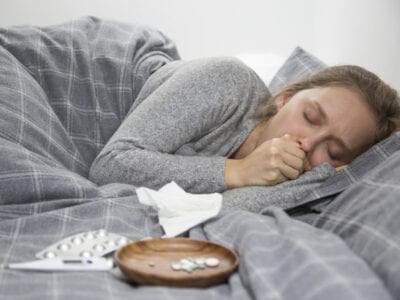 Photo of sick young woman in bed coughing with thermometer and medicine beside her.